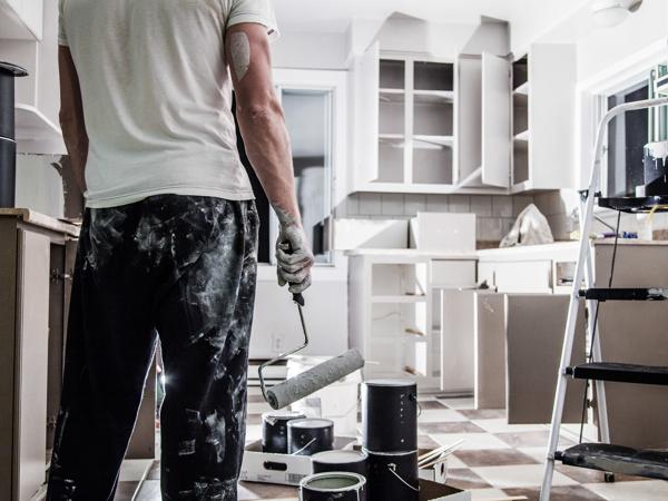 A man holding a paint roller in a kitchen mid-renovation, with the upper cabinet doors off